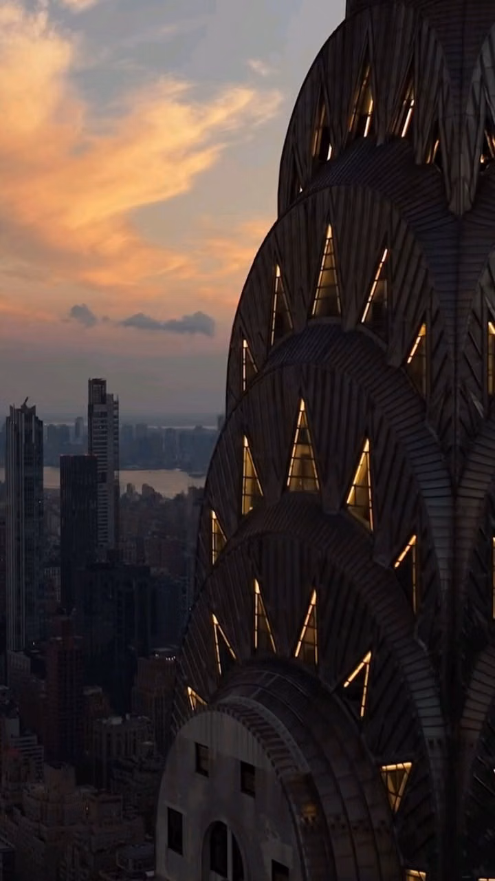 Chrysler Building crown close up photograph - The iconic Chrysler Building crown illuminated at golden hour, its stainless steel Art Deco eagle arches and triangular windows glowing against a dramatic New York City sunset. One of the most breathtaking architectural views in Manhattan, captured from above with the Hudson River and city skyline stretching into the distance. A must-see landmark for architecture lovers, NYC travel enthusiasts, and urban photography fans.