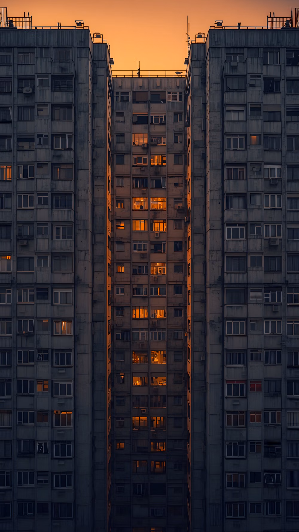 Brutalism aesthetic - Symmetrical view of a residential building at dusk. Warm lights glow in the windows against the dark concrete. A moody look at city life and modern architecture. High-quality vertical aesthetic for your lockscreen. brutalist architecture, urban windows, city life aesthetic, apartment building light, moody city, dusk photography, symmetrical building, ventanas de la ciudad, arquitectura brutalista, luces nocturnas, edificios de apartamentos, estética urbana, fondos de pantalla oscuros. #urbanlights #brutalism #citywindows #apartmentliving #moodycity #architecturephotography #nightaesthetic #cityscape #fondosdepantalla #visualsatisfaction #modernbuilding #urbanview